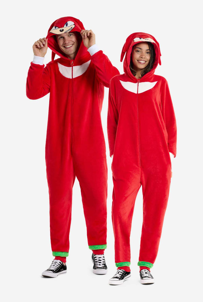 Ein Mann und eine Frau tragen einen leuchtend roten Knuckles the Echidna-Einteiler für Erwachsene von OppoSuits. Der lustige und kuschelige Einteiler verfügt über einen Reißverschluss vorne und eine angenähte Kapuze, die dem Gesicht von Knuckles nachempfunden ist, komplett mit ausdrucksstarken Augen und schlaffen Dreadlocks. Der Anzug ist hauptsächlich rot, mit einem weißen Halbmondmuster auf der Brust, das Knuckles' ikonischen Look aus der Videospielserie Sonic the Hedgehog widerspiegelt. Full body image