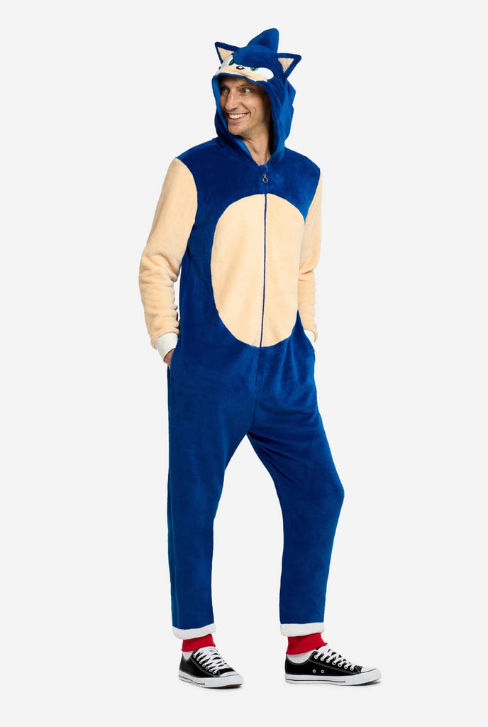 A man is wearing a playful blue and beige Sonic the Hedgehog-themed adult onesie from OppoSuits. The cozy onesie features a zip-up front and an attached hood designed to resemble Sonic’s iconic face, complete with ears and an expressive gaze. The blue body of the suit contrasts with a beige belly, mimicking Sonic's classic look. Holding his hands in his pocket.