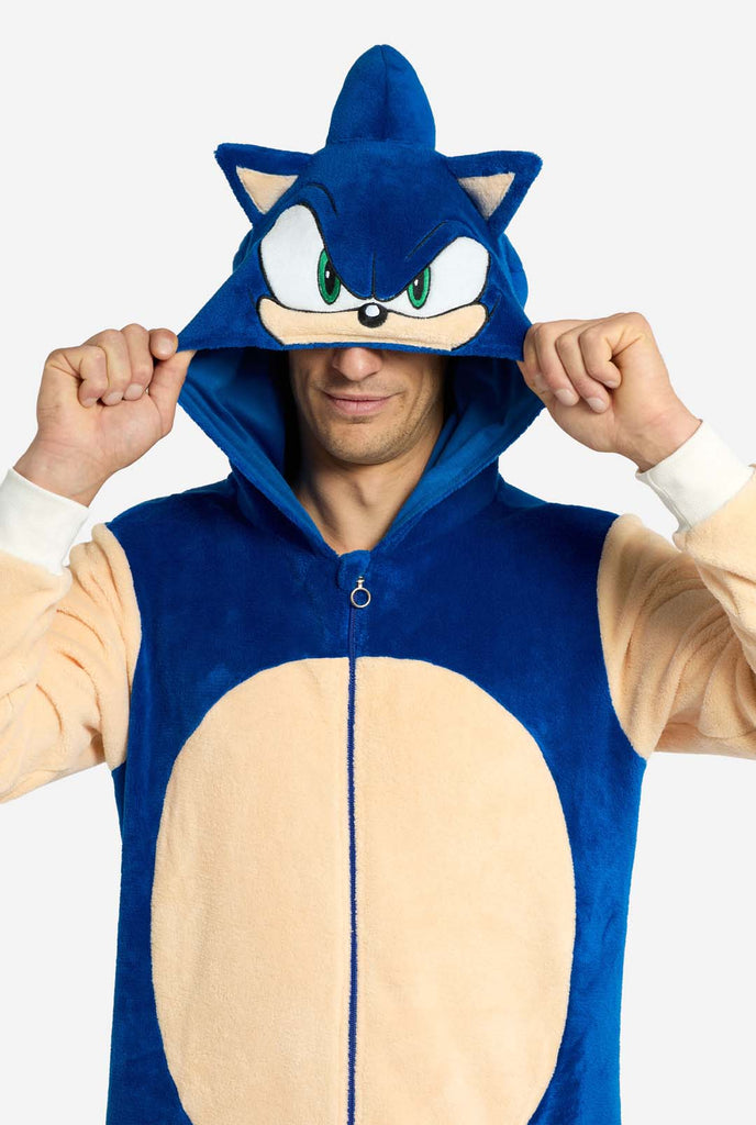 A man is wearing a playful blue and beige Sonic the Hedgehog-themed adult onesie from OppoSuits. The cozy onesie features a zip-up front and an attached hood designed to resemble Sonic’s iconic face, complete with ears and an expressive gaze. The blue body of the suit contrasts with a beige belly, mimicking Sonic's classic look. Holding the hood over his eyes.