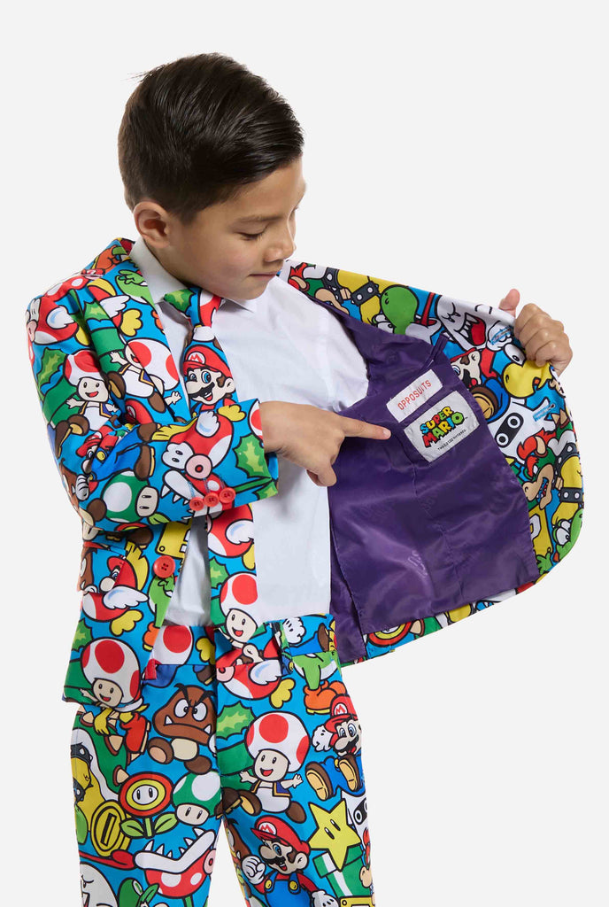 Bild eines Kindes, das einen Anzug mit Super Mario-Allover-Print trägt, bestehend aus Blazer, Hose und Krawatte. Die Hauptfarbe ist blau, aber es gibt auch gelb, rot und weiß.