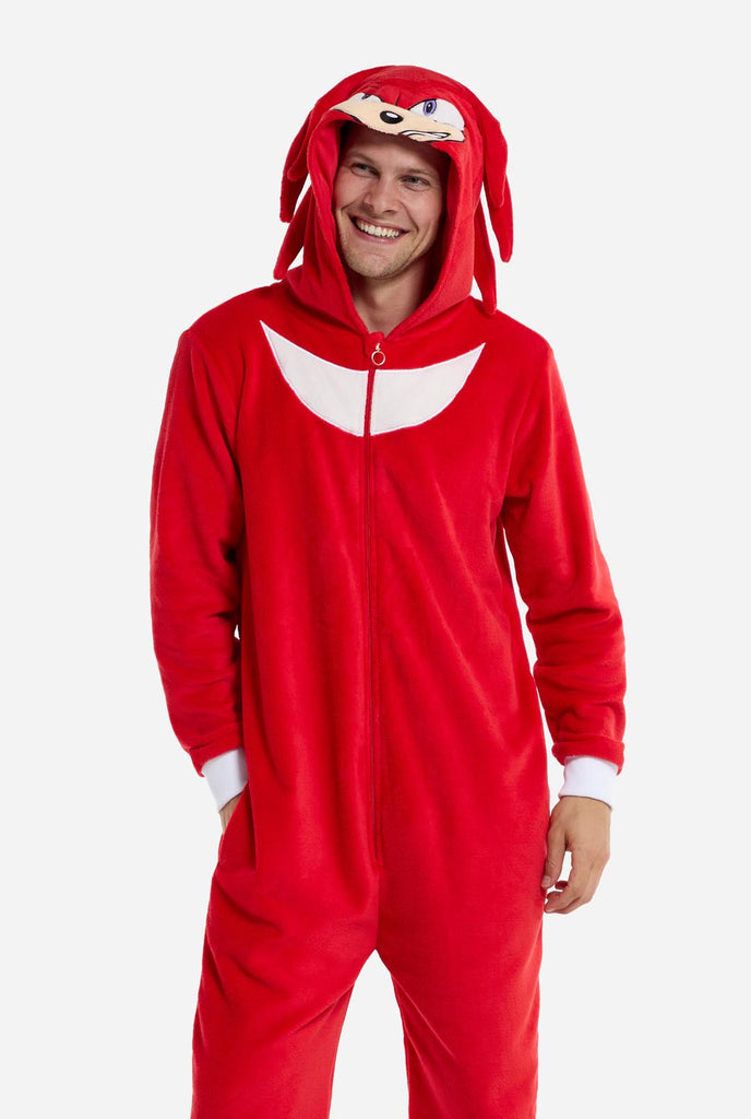 Ein Mann trägt einen leuchtend roten Knuckles the Echidna-Einteiler für Erwachsene von OppoSuits. Der lustige und kuschelige Einteiler verfügt über einen Reißverschluss vorne und eine angenähte Kapuze, die dem Gesicht von Knuckles nachempfunden ist, komplett mit ausdrucksstarken Augen und schlaffen Dreadlocks. Der Anzug ist hauptsächlich rot, mit einem weißen Halbmondmuster auf der Brust, das Knuckles' ikonischen Look aus der Videospielserie Sonic the Hedgehog widerspiegelt.