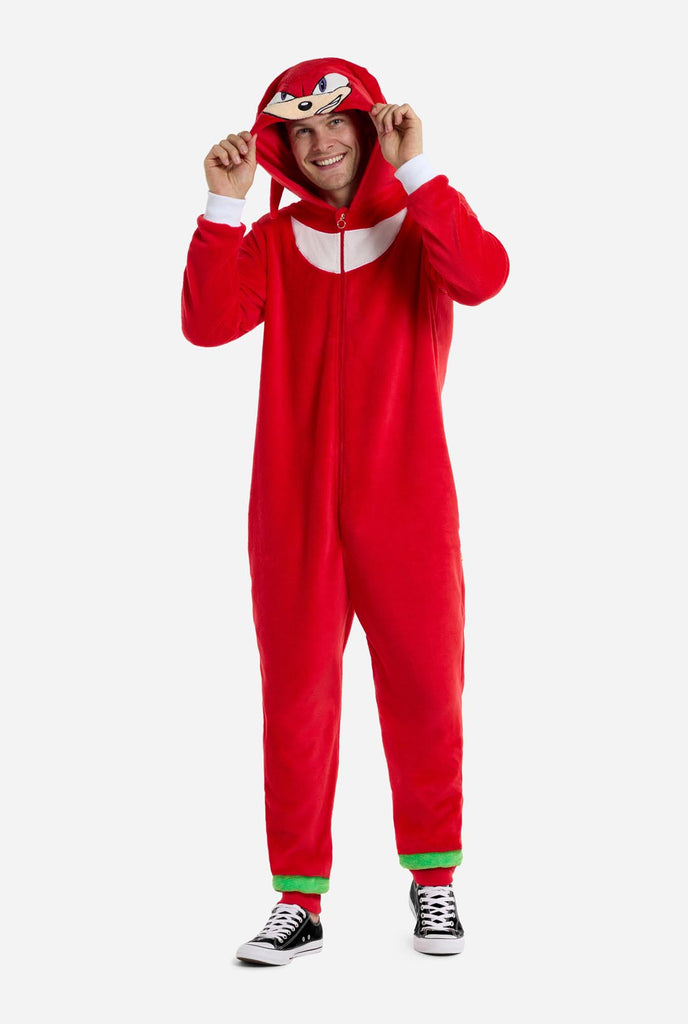 Ein Mann trägt einen leuchtend roten Knuckles the Echidna-Einteiler für Erwachsene von OppoSuits. Der lustige und kuschelige Einteiler verfügt über einen Reißverschluss vorne und eine angenähte Kapuze, die dem Gesicht von Knuckles nachempfunden ist, komplett mit ausdrucksstarken Augen und schlaffen Dreadlocks. Der Anzug ist hauptsächlich rot, mit einem weißen Halbmondmuster auf der Brust, das Knuckles' ikonischen Look aus der Videospielserie Sonic the Hedgehog widerspiegelt.