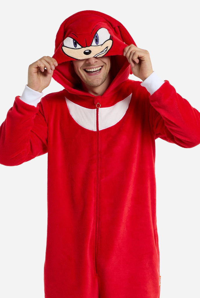 Ein Mann trägt einen leuchtend roten Knuckles the Echidna-Einteiler für Erwachsene von OppoSuits. Der lustige und kuschelige Einteiler verfügt über einen Reißverschluss vorne und eine angenähte Kapuze, die dem Gesicht von Knuckles nachempfunden ist, komplett mit ausdrucksstarken Augen und schlaffen Dreadlocks. Der Anzug ist hauptsächlich rot, mit einem weißen Halbmondmuster auf der Brust, das Knuckles' ikonischen Look aus der Videospielserie Sonic the Hedgehog widerspiegelt.