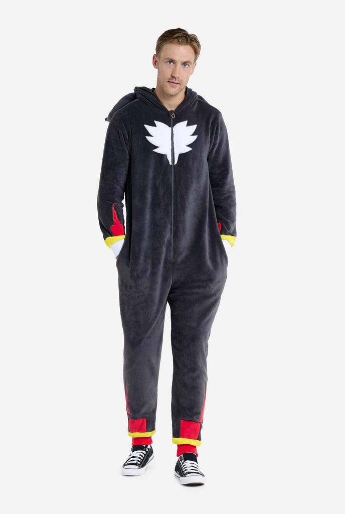 Ein Mann trägt einen plüschigen schwarzen Onesie, der von der beliebten Igelfigur Shadow inspiriert ist, mit stacheligen roten und schwarzen Akzenten auf der Kapuze, einem weißen Brustemblem und gelben Handgelenkdetails. Der Onesie ist aus weichem Fleecematerial gefertigt und bietet Komfort und ein verspieltes Design für Cosplay, Relaxen oder Themenveranstaltungen.