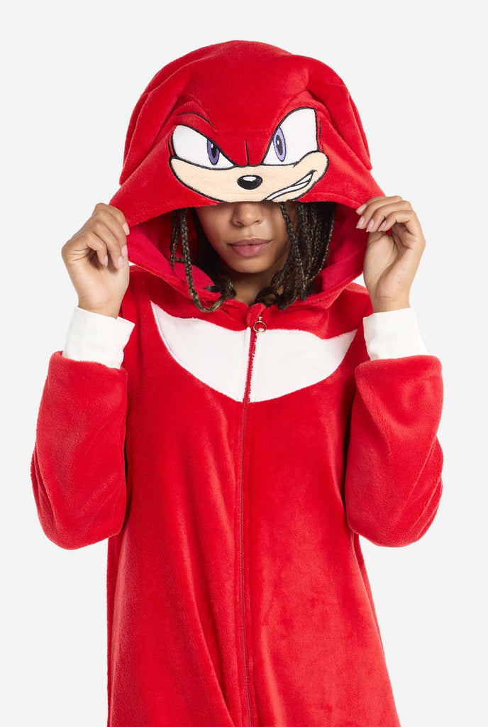 Eine Frau trägt einen leuchtend roten Knuckles the Echidna-Einteiler für Erwachsene, ein gemütliches und verspieltes Outfit, das von der berühmten Figur aus der Sonic the Hedgehog-Serie inspiriert ist. Der Einteiler ist mit dem charakteristischen weißen Aufnäher von Knuckles auf der Brust versehen und verfügt über eine Kapuze, die Knuckles‘ Gesicht mit seinen ausdrucksstarken Augen und langen roten Dreadlocks zur Geltung bringt. Der vordere Reißverschluss sorgt für eine bequeme Passform.