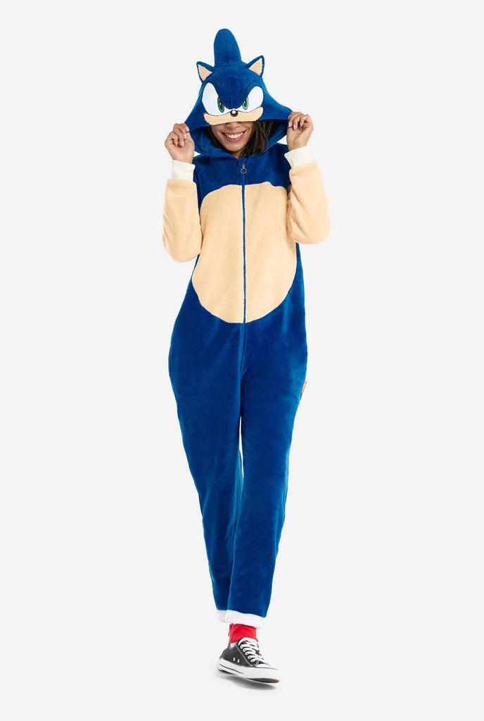 A woman is dressed in a cozy, blue Sonic the Hedgehog adult onesie, featuring soft beige patches on the front and sleeves to replicate Sonic's iconic look. The onesie has a hood designed with Sonic's recognizable face, including large green eyes, blue spikes, and pointy ears. The front zipper closure ensures a comfortable fit, and the woman is holding the hood over her eyes. Her expression is calm and relaxed. This Sonic the Hedgehog onesie is ideal for cosplay, lounging, or themed events. Front view.